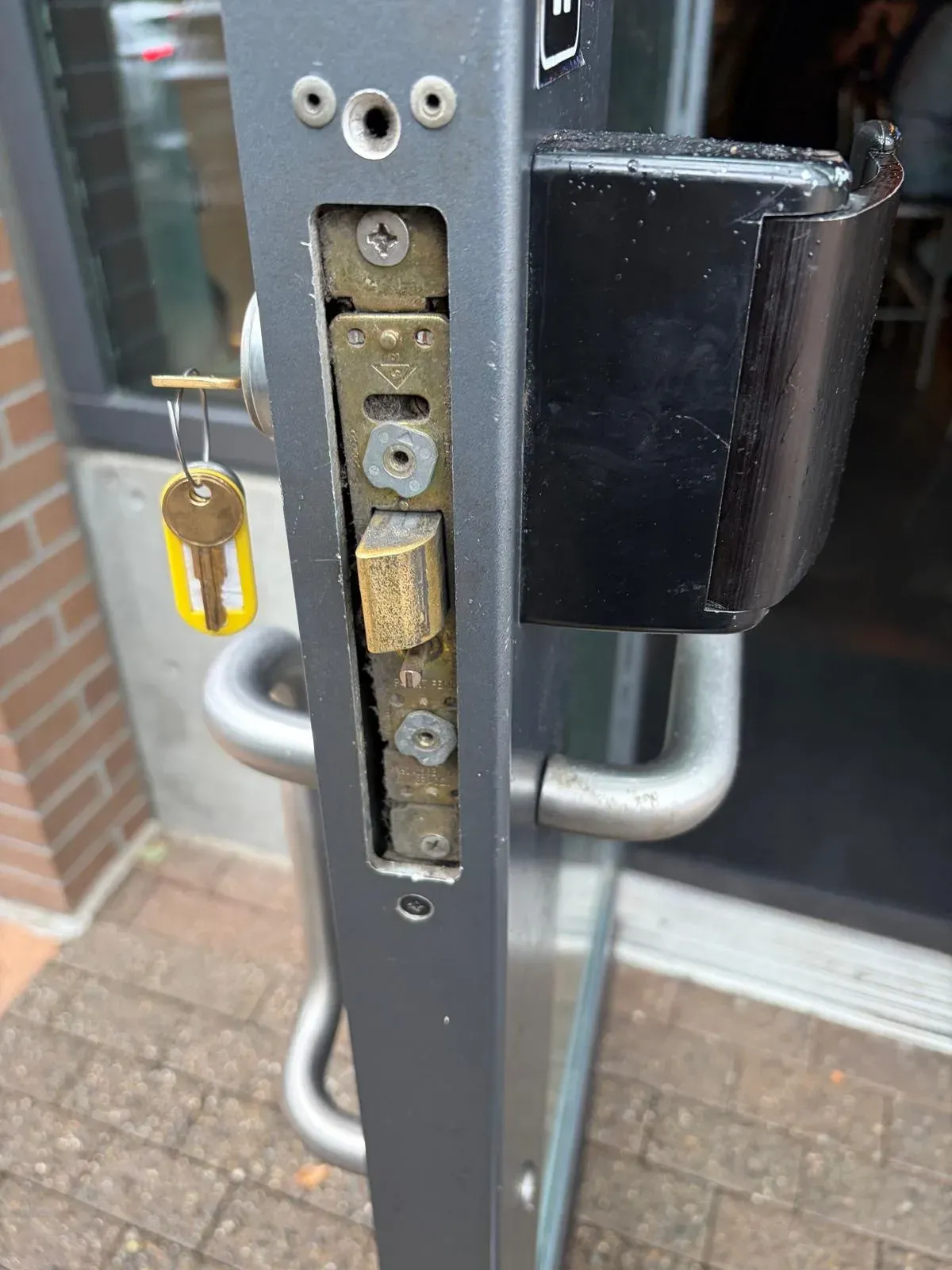 Open storefront door with its mortise lock exposed and a key with a yellow tag